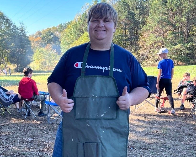 Smiling person in green apron giving thumbs up outdoors with camping chairs and trees behind them