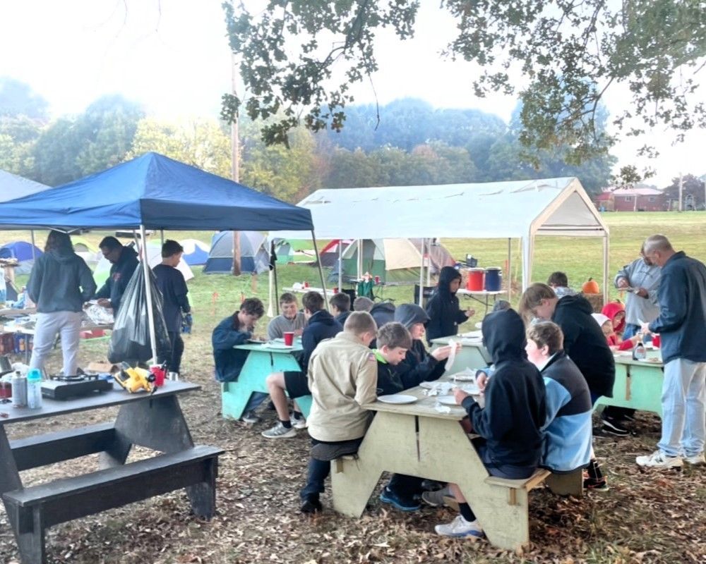 People gathered at picnic tables under tents in a park for an outdoor event.