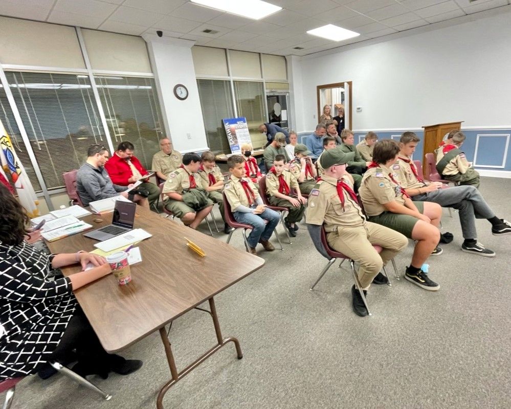 Scout troop seated in a meeting room, listening to a speaker at a table.