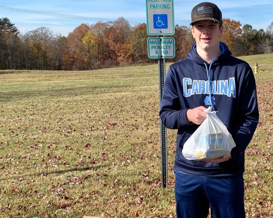 Person in a Carolina sweatshirt holding a takeout bag beside a handicap parking sign in a grassy lot.