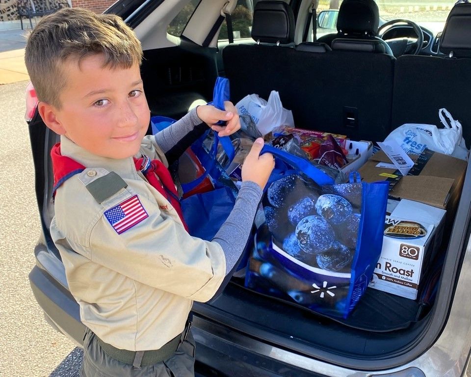 Boy in a car trunk packing a blue bag of supplies and groceries, smiling toward the camera.