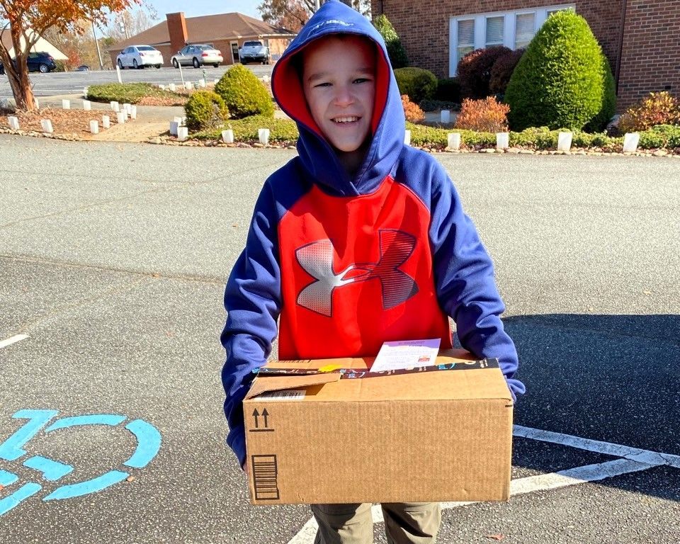 Child in a blue-and-red hoodie holding a cardboard box in a parking lot outdoors