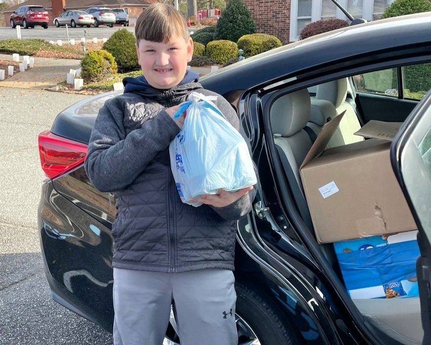 Person holding a blue bag beside an open car trunk with boxes and supplies.