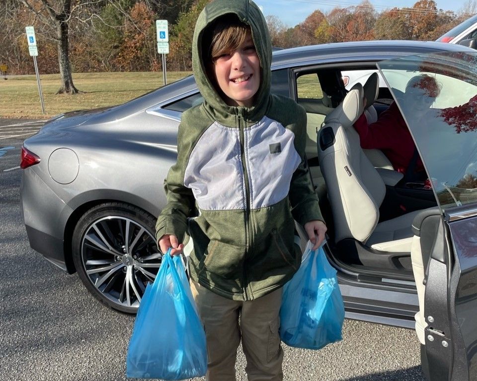 Person in a green jacket stands by an open car door, holding two blue plastic bags.