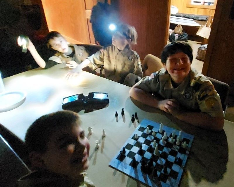 Children playing chess at a table, one smiling at the camera, in a dimly lit room.