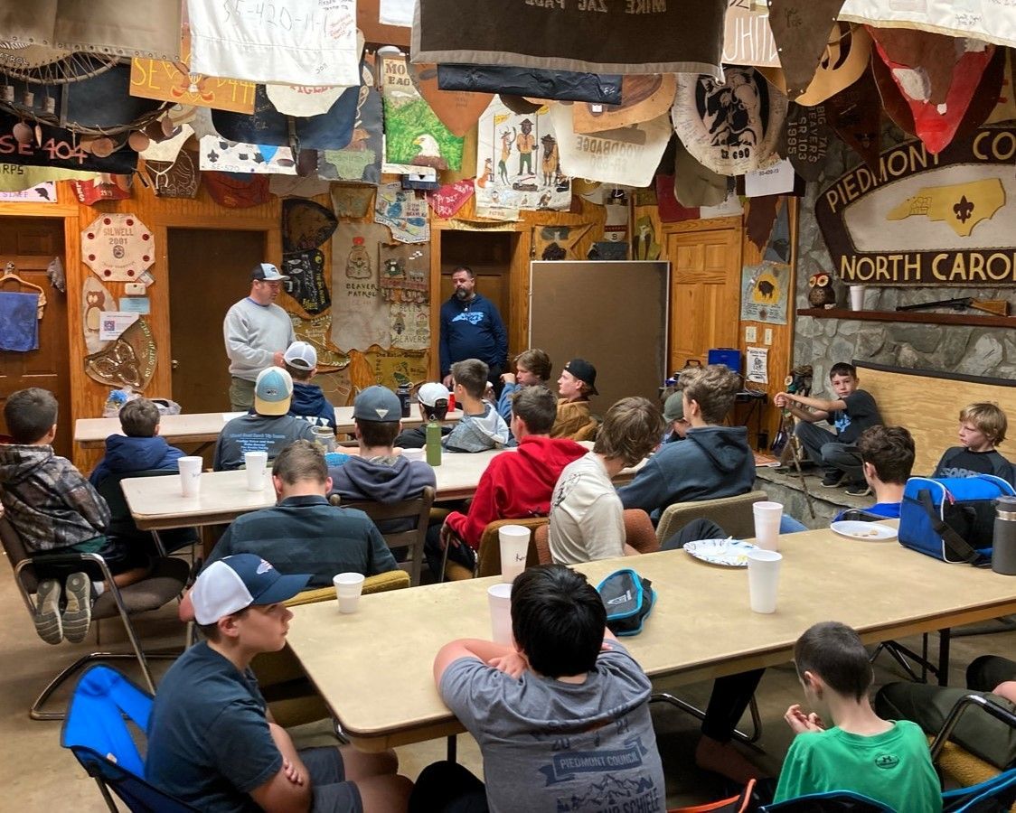 Children seated at tables watching a man speak in a rustic classroom or museum room