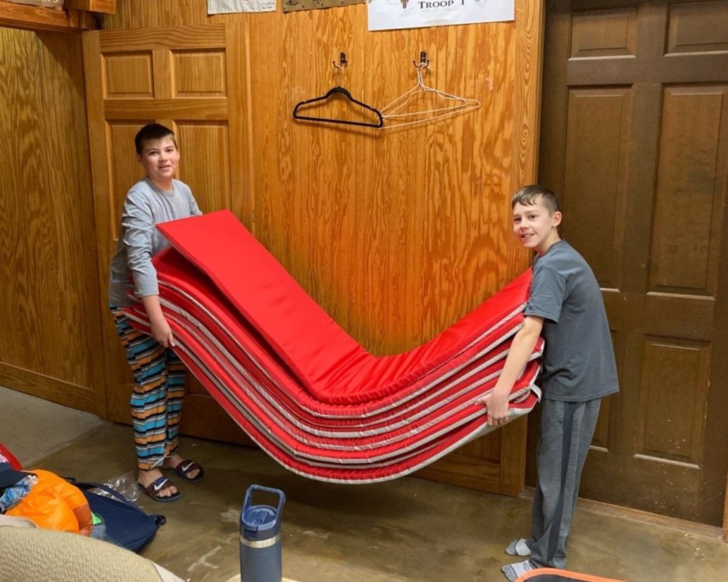 Two boys carrying a stack of red mats in a wood-paneled room