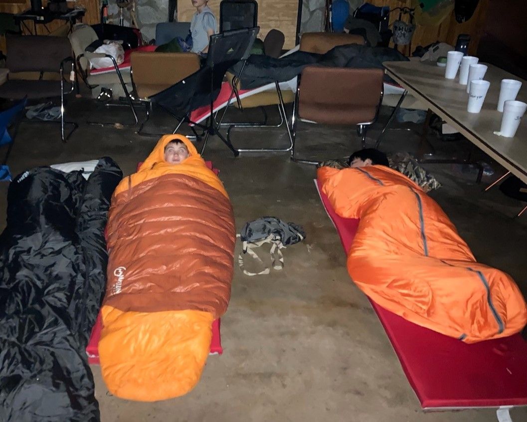 Three sleeping bags on the floor of a cluttered room, with folding chairs and cups in the background.