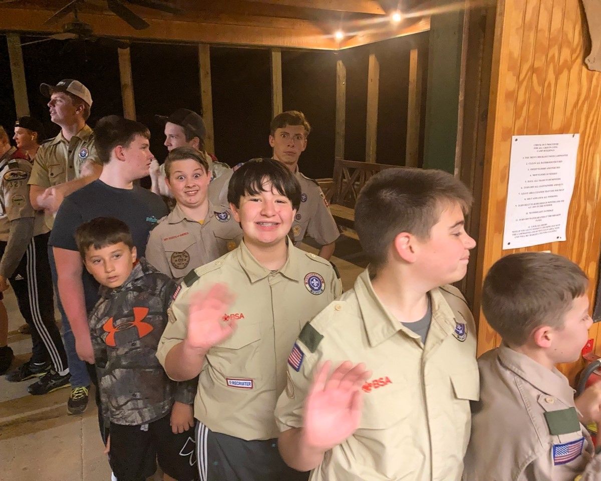 Boy Scouts in tan uniforms stand indoors, smiling and waving in a wooden cabin.