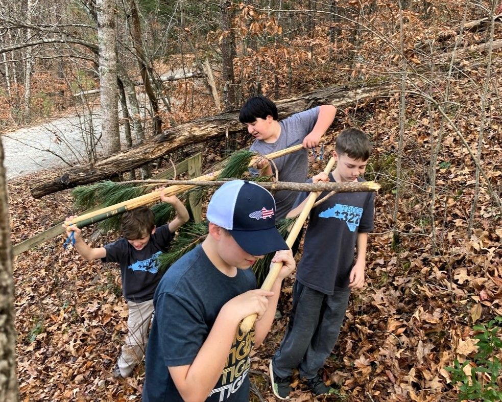 Four hikers carrying logs through a wooded, leaf-covered trail