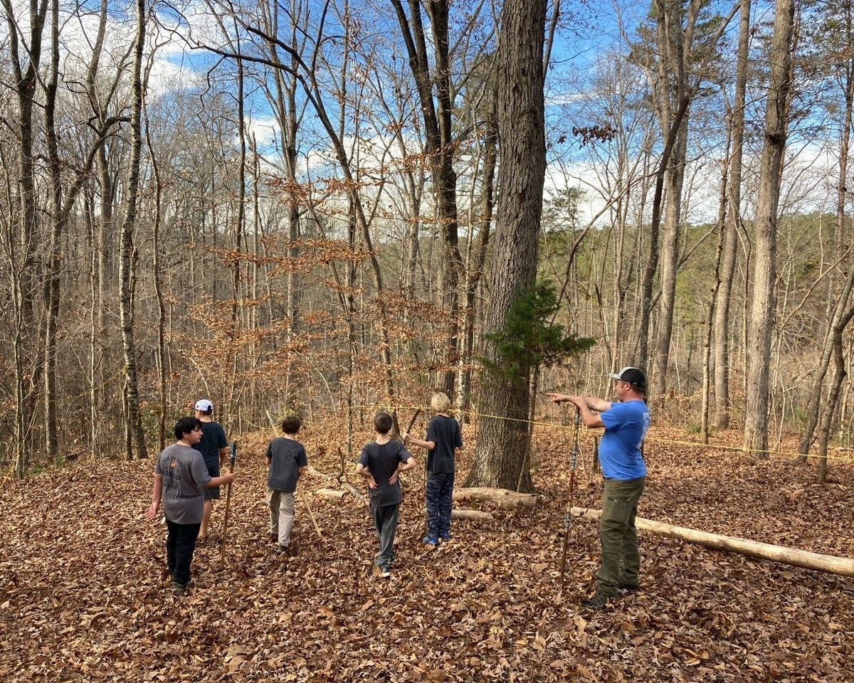 Five people stand in a wooded area, one pointing while others listen near a fallen log.