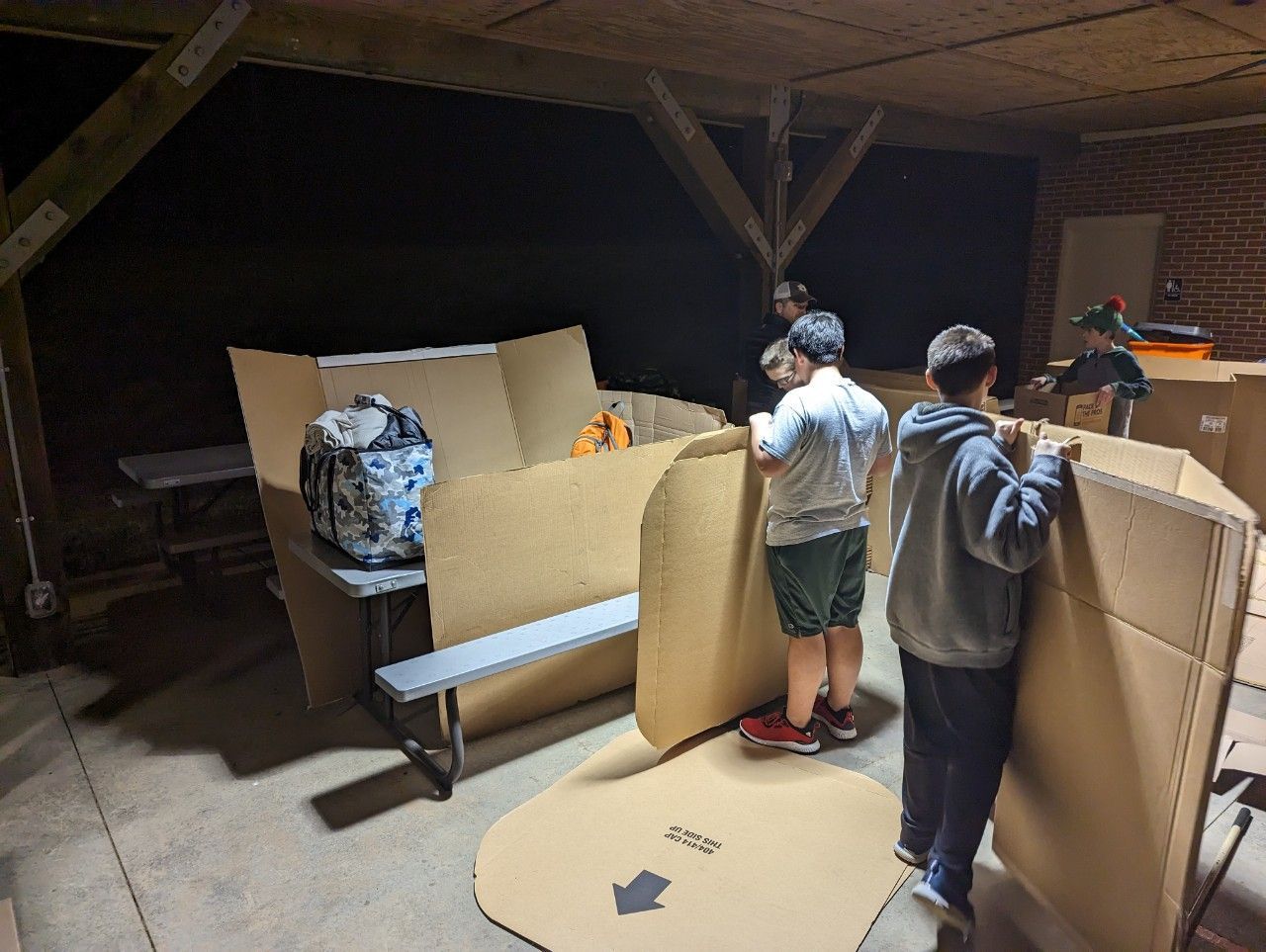 Two children building a cardboard fort with panels and a bench in a dim attic room.