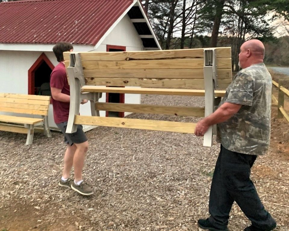 Two people carry a wooden bench outdoors near a red-roofed building.