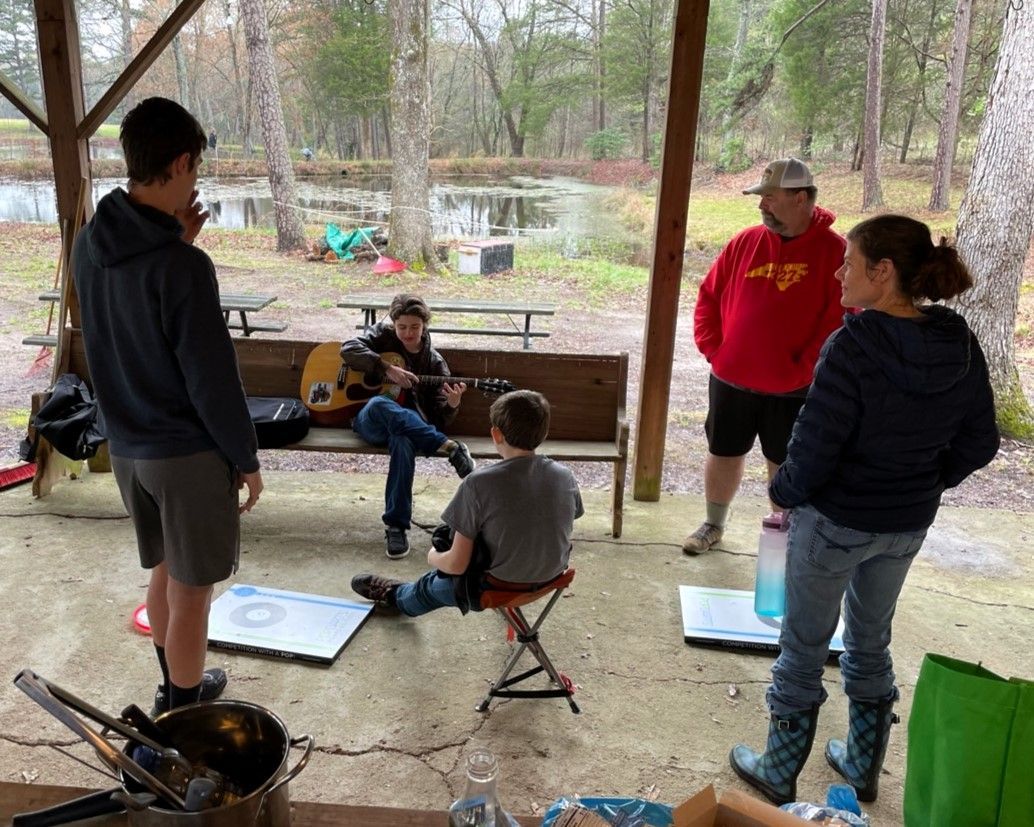People gathered around a campfire at a wooded picnic shelter near a river, with chairs and picnic tables nearby.