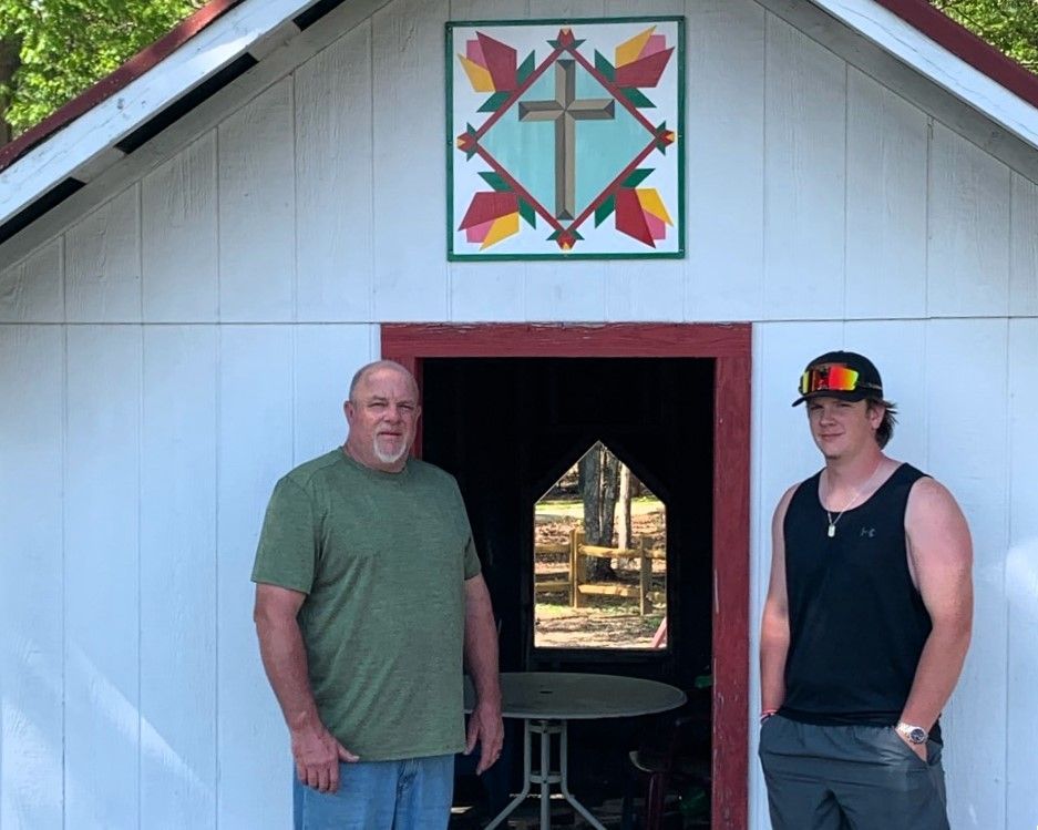 Two people standing in a white pavilion doorway beneath a colorful quilt square decoration.