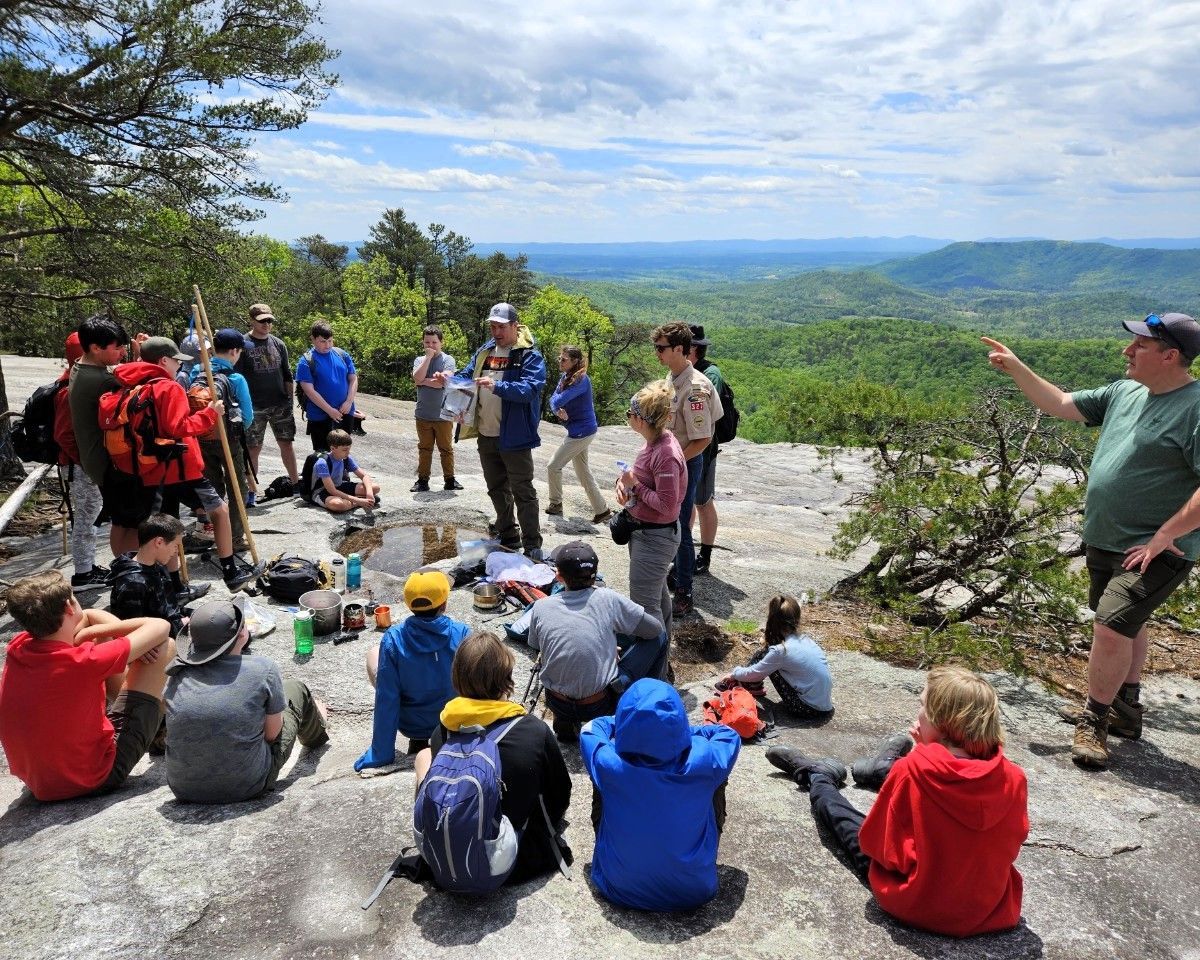 Group of hikers resting on a rocky overlook with backpacks, overlooking a forested mountain valley