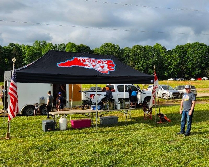 Tailgate tent with black canopy and red logo, flags, vehicles, and a man standing on a grassy field.