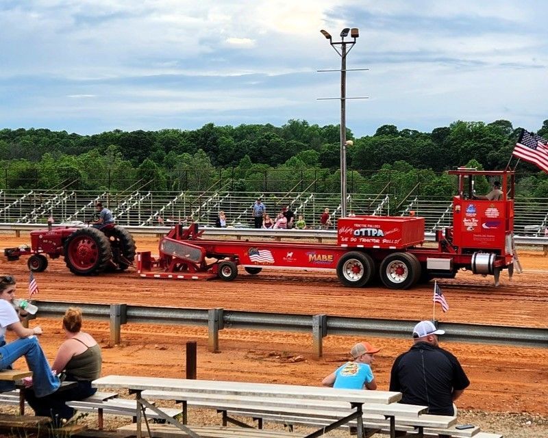 Red tractor pulling a long trailer on a dirt track at a fairground, with spectators in bleachers.