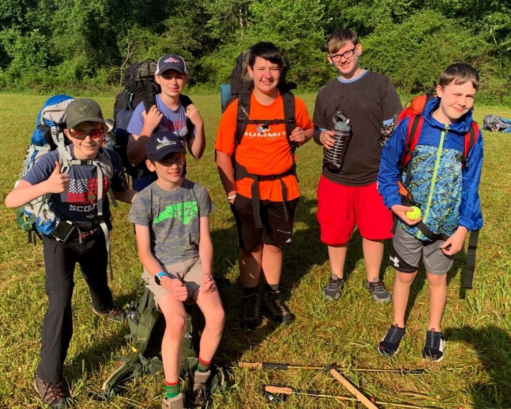 Six hikers with backpacks standing on a grassy trail in bright outdoor sunlight
