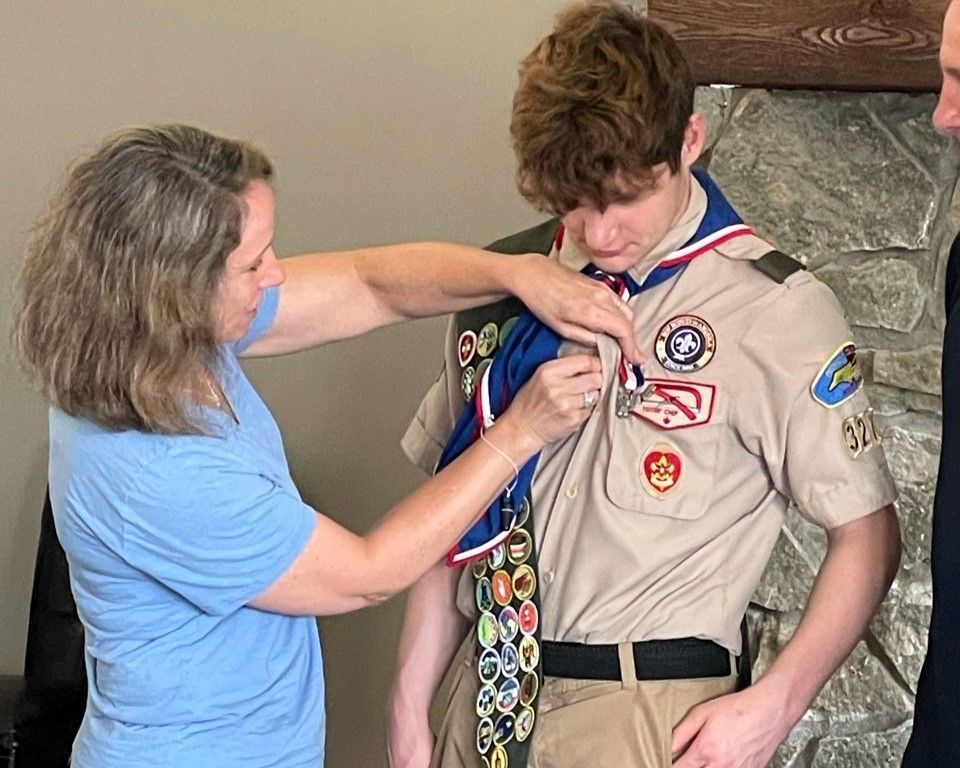 Woman in blue shirt adjusting a scout neckerchief on a uniformed youth indoors