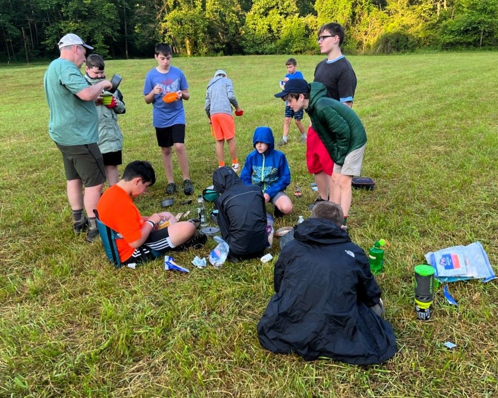 Group of people outdoors on a grassy field, gathered around camping gear and cooking equipment.