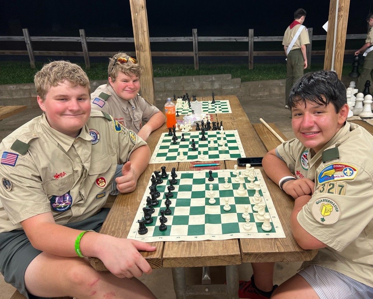 Three boy scouts are sitting at a picnic table playing chess.