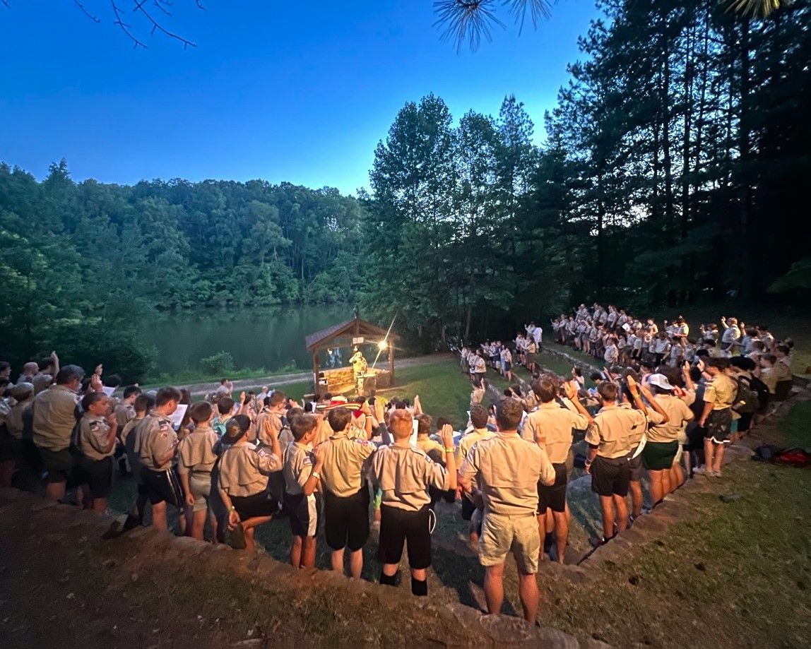A large group of people are standing in a circle in front of a lake.