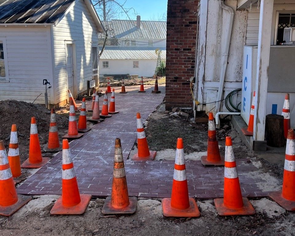 Orange traffic cones line a narrow construction path beside a house and brick wall