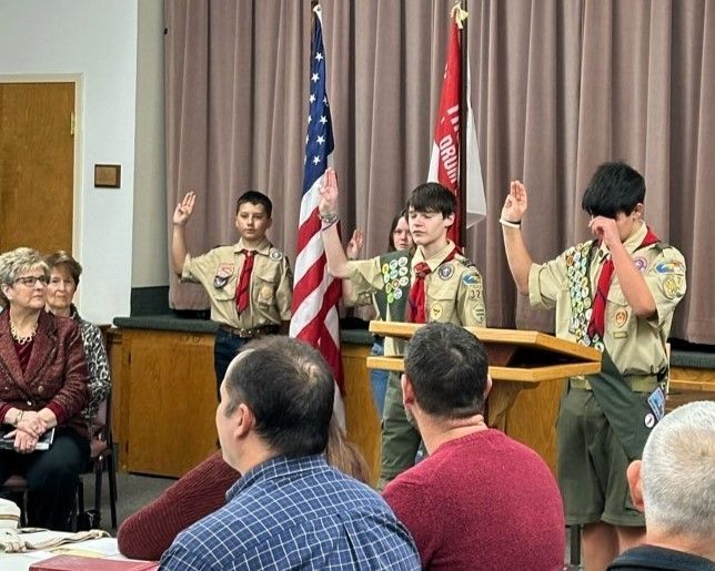 Three Scouts in uniform raise right hands during a ceremony indoors, with flags behind them and an audience seated nearby.