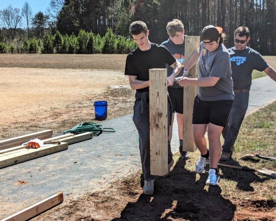 Four people outdoors working together to stand up a wooden post in a dirt area
