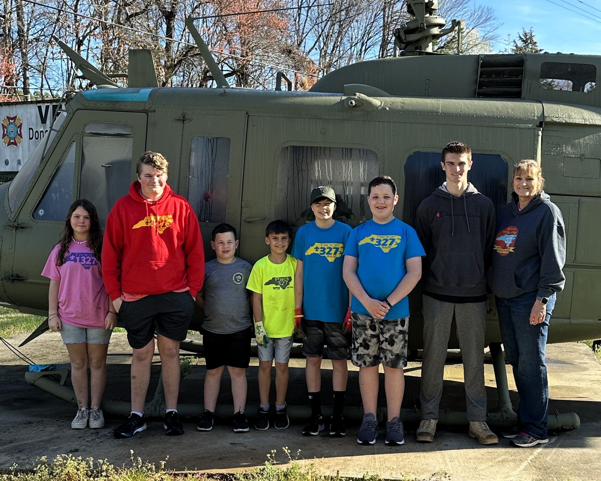 Seven people posing in front of a military helicopter outdoors on a sunny day.