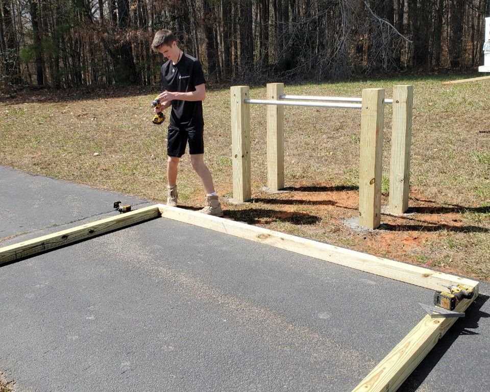 Person stands beside a wooden barrier across a paved path in a wooded area.