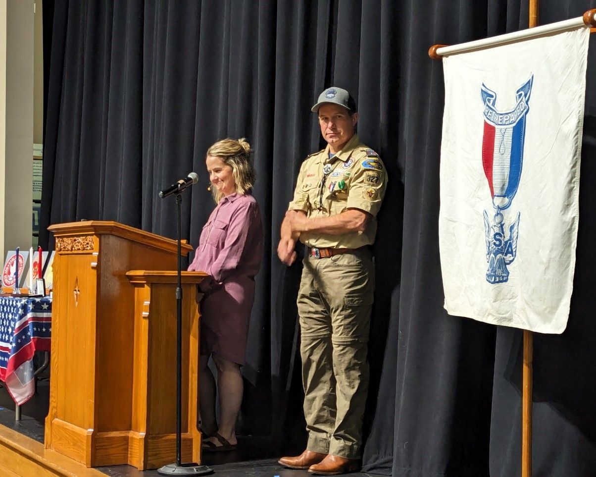 Two people on a stage beside a podium and microphone, with a hanging banner backdrop.