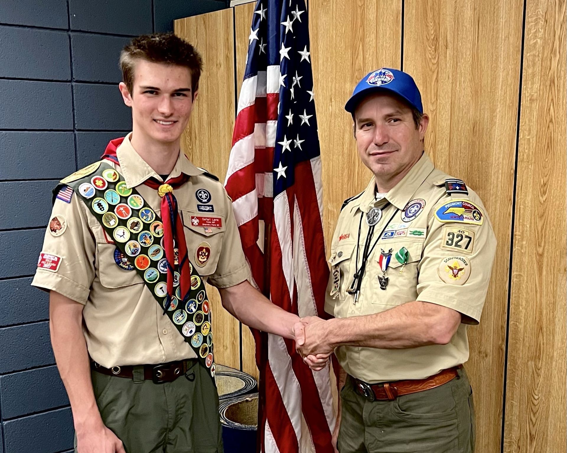 Two Scouts in tan uniforms holding an American flag indoors, standing side by side and shaking hands