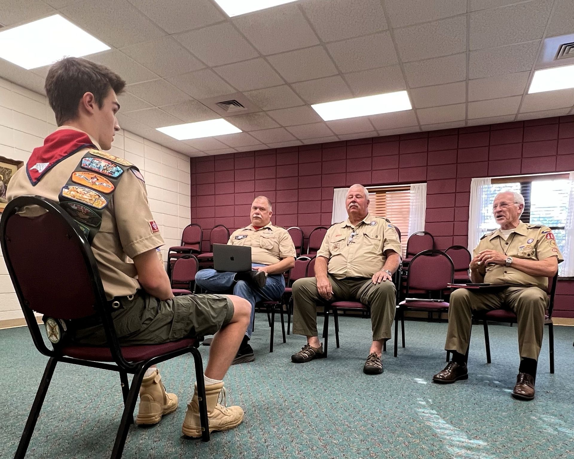 Boy Scouts meeting with three uniformed leaders in a room, seated in a circle with a laptop open.