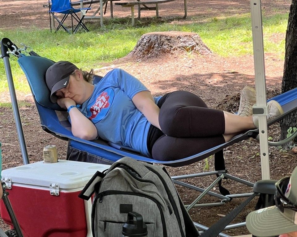 Person sleeping in a blue camp chair under a canopy, with bags and cooler nearby outdoors.