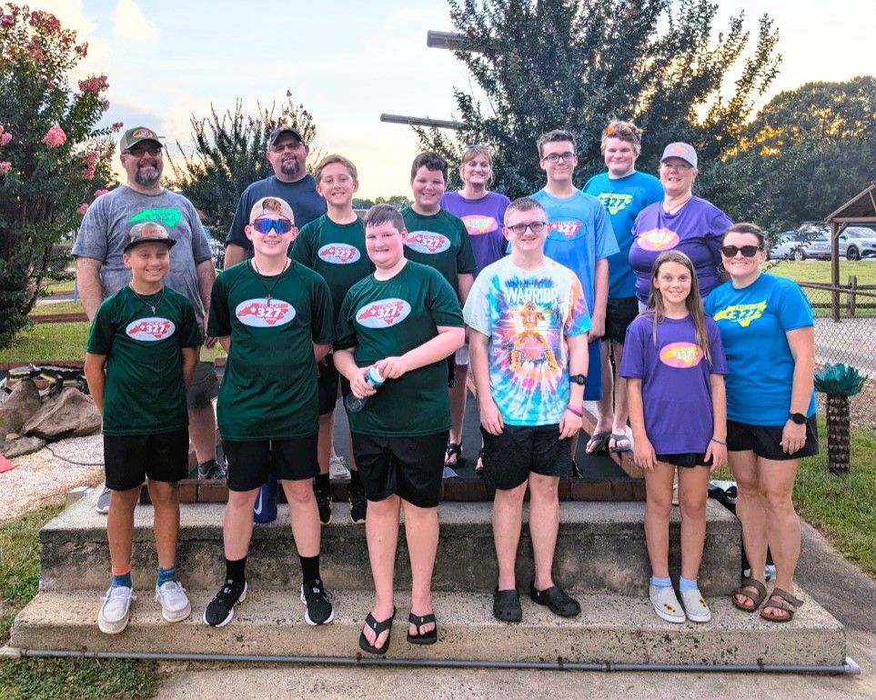 Large group posing outdoors at dusk, wearing matching shirts in front of a landscaped garden.