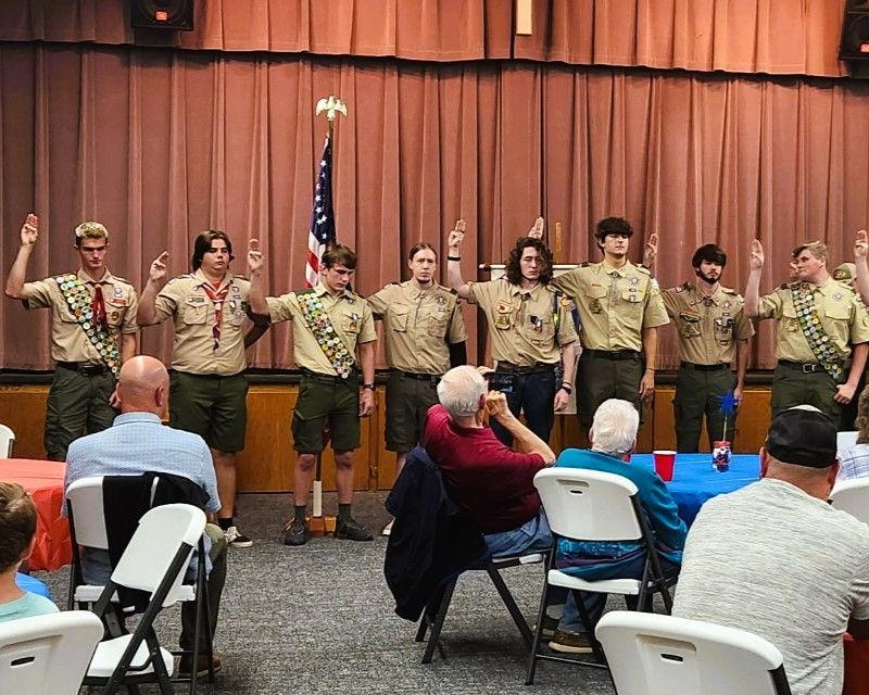 Scouts in tan uniforms stand on a stage, saluting in front of a curtain and American flag while an audience watches.