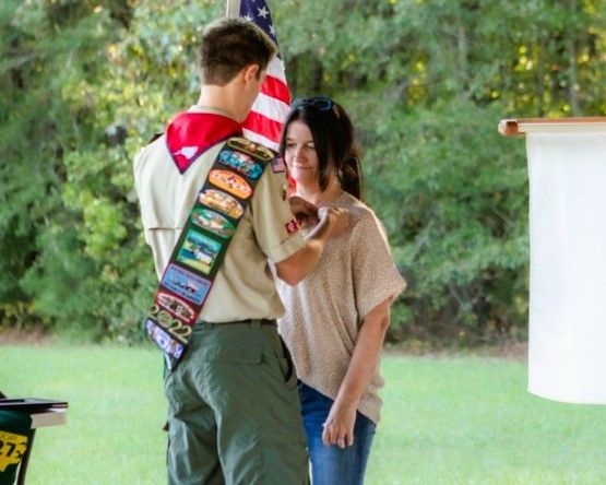 Two people stand outdoors; one wears a Scout uniform with merit badges and an American flag scarf.