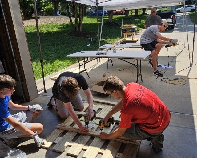 People building a wooden pallet structure outdoors beside a sidewalk and picnic tables.