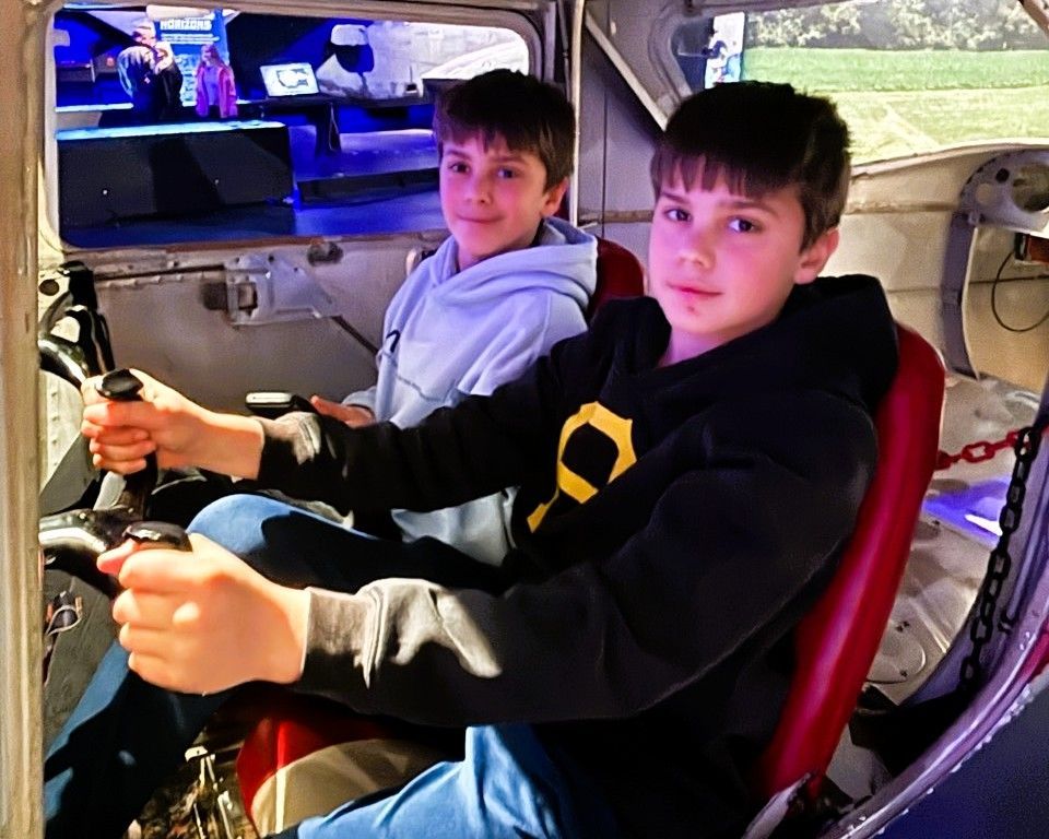 Two boys sitting in a vehicle cockpit, holding controls and smiling, with a grassy landscape outside.