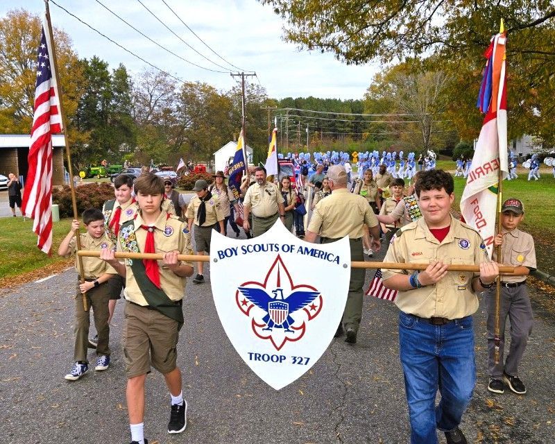 Boy Scouts march in a parade carrying a Troop 727 banner and flags on a tree-lined road.