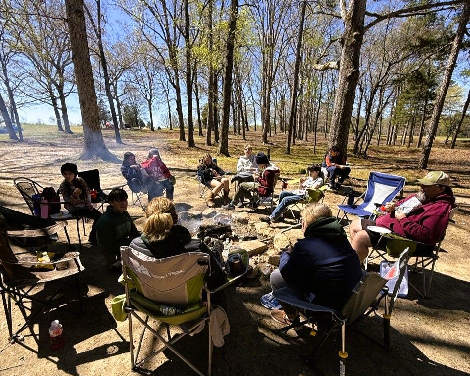 People sitting in folding chairs around a campfire in a wooded park on a sunny day