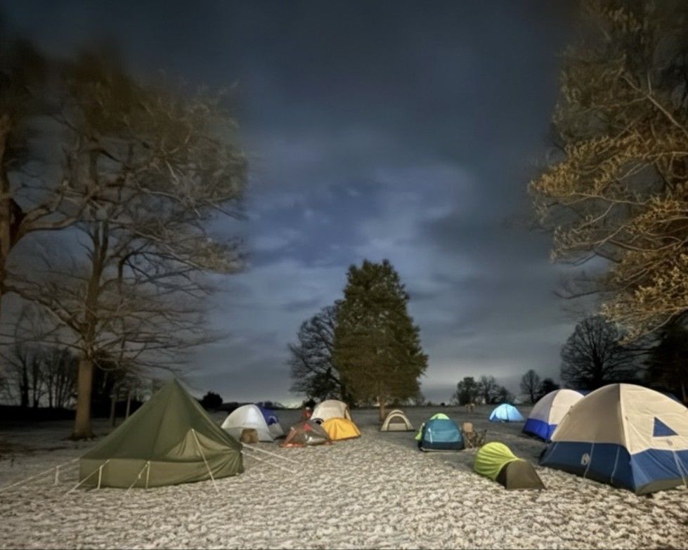 Tents set up on a gravel campsite at night under a cloudy sky, with trees framing the scene.