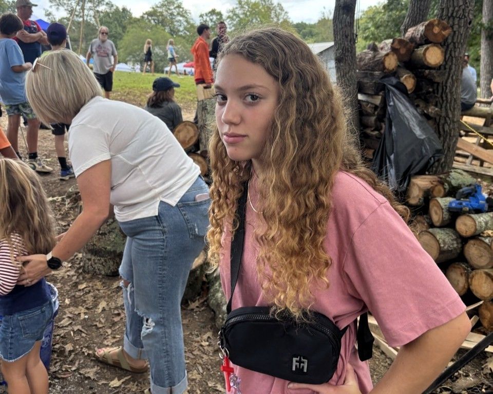 Teen in pink shirt stands outdoors near a woodpile at a gathering, looking at the camera.