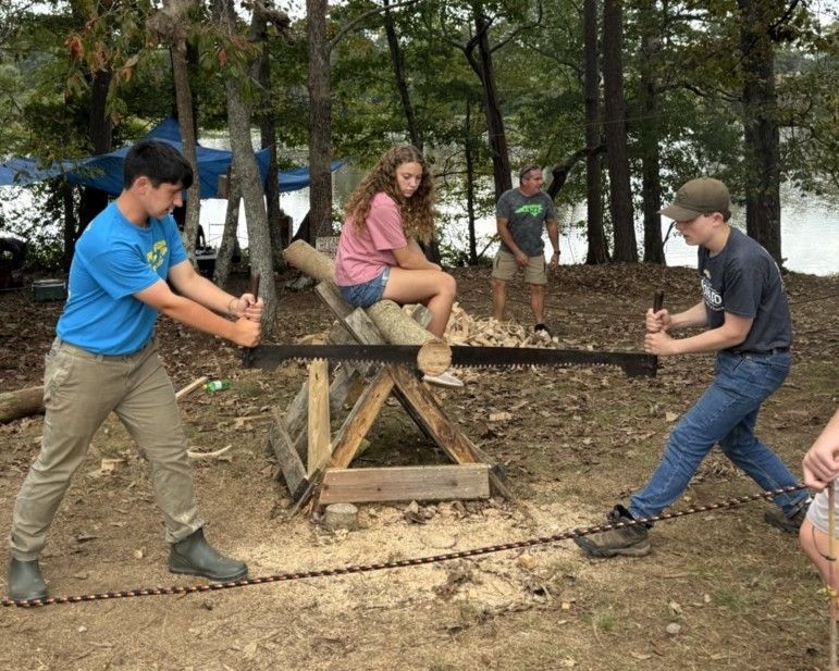 People outdoors using a wooden seesaw-like device in a wooded area, with two others watching nearby.