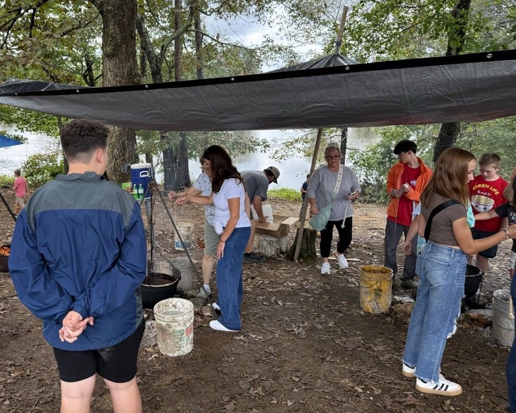 People gathered under a canopy outdoors, cooking and chatting around buckets and a fire pit.