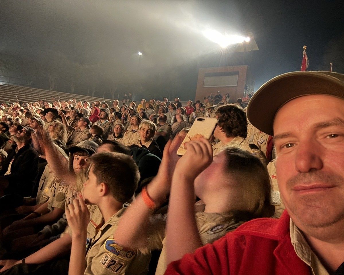 A man in a red shirt is taking a selfie in front of a crowd of people