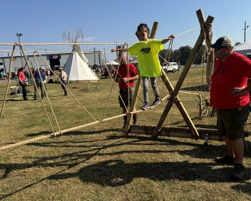 A boy in a yellow shirt is standing on a wooden rope bridge.