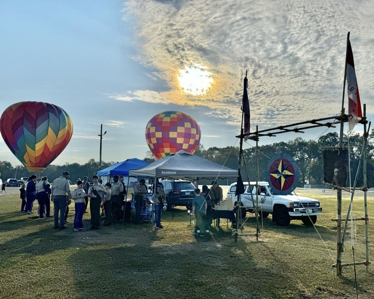 A group of people are standing in a field with hot air balloons in the background.